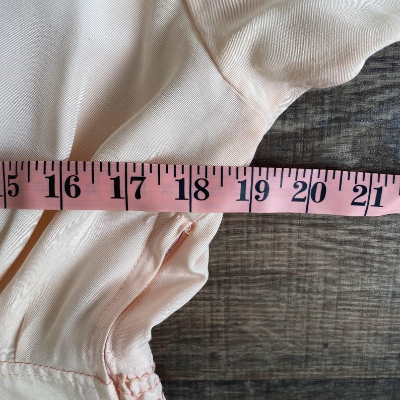 May include: A light pink garment featuring a side seam and a pink measuring tape placed horizontally across the fabric. The measuring tape clearly displays numerical markings from 15 to 21 inches, indicating the width of the garment section.