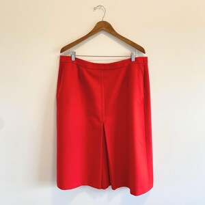 May include: A bright red knee-length skirt hanging on a wooden hanger. The skirt features a structured waistband, a clean front design, and a prominent vertical center pleat that extends from the waist down to the hemline, creating a tailored look.
