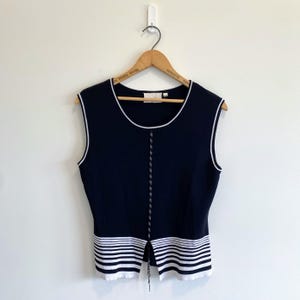 May include: A sleeveless navy blue knit top featuring a round neckline with white trim. The garment includes a vertical center stripe detail and a horizontal striped pattern at the hem, which is split in the middle, hanging on a wooden hanger.