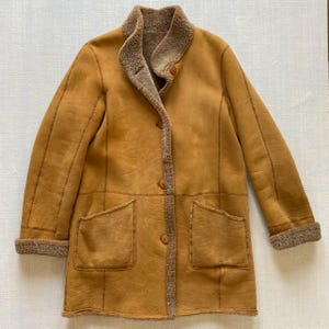Puede incluir: Un abrigo de color tostado con un exterior liso y un forro texturizado y difuso visible en el cuello, los puños y la abertura frontal. El abrigo cuenta con mangas largas, dos grandes bolsillos de parche en la parte delantera y tres botones.