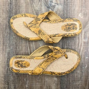May include: A pair of thong sandals featuring a yellow and black snakeskin pattern on the straps and footbed edges. The beige footbed is embossed with a floral pattern and the b.o.c. brand logo is printed on the heel area of each sandal.