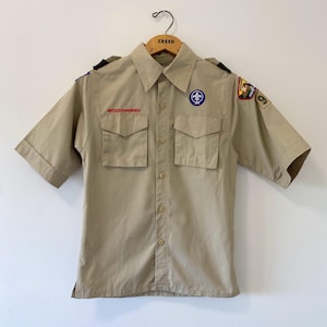 May include: A tan short-sleeved button-down shirt featuring two chest pockets, a collar, and shoulder epaulets. The shirt displays the text Boys Scouts of America, a blue fleur-de-lis emblem, and various embroidered patches on the sleeves.
