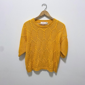 May include: A mustard yellow short-sleeve sweater featuring a prominent cable knit pattern across the front. The garment has a round neckline, ribbed trim at the collar, cuffs, and hem, and is displayed on a wooden hanger with the text Park Plaza Hotel Toronto.