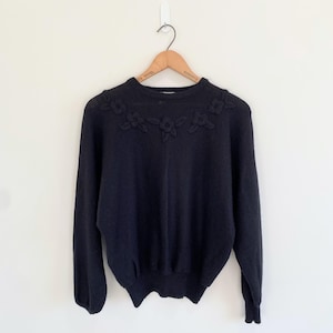May include: A black long-sleeve sweater featuring a crew neckline and a decorative floral knit pattern across the chest. The garment is displayed on a wooden hanger, showcasing a ribbed collar, cuffs, and hem with a solid black knit texture.