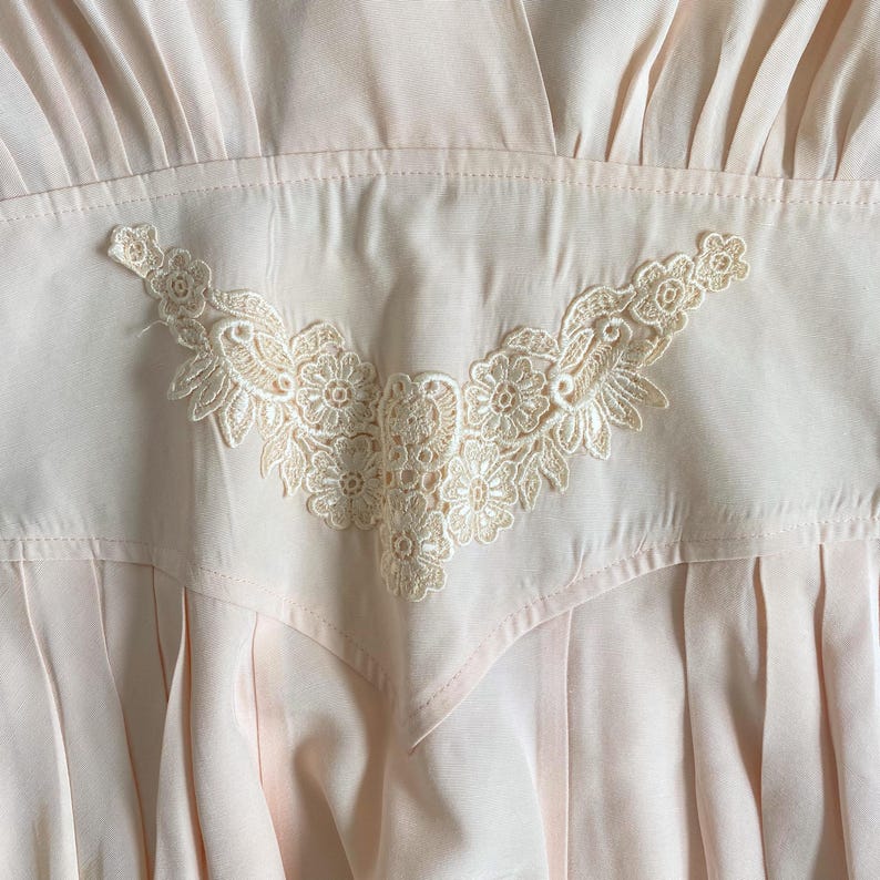 May include: A pale pink dress featuring a V-shaped bodice panel adorned with intricate cream-colored floral lace embroidery. The garment includes a gathered waistline and vertical pleats extending downward from the lace-detailed midsection.