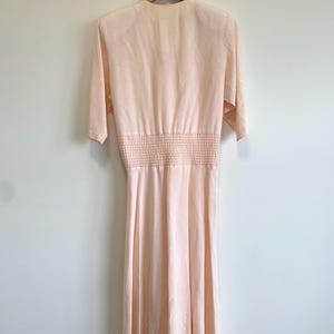 May include: A pale peach-colored midi dress featuring a V-neckline, elbow-length sleeves, and a wide smocked elasticized waist panel. The garment has a smooth, flowing skirt that falls below the knees and is displayed on a wooden hanger.