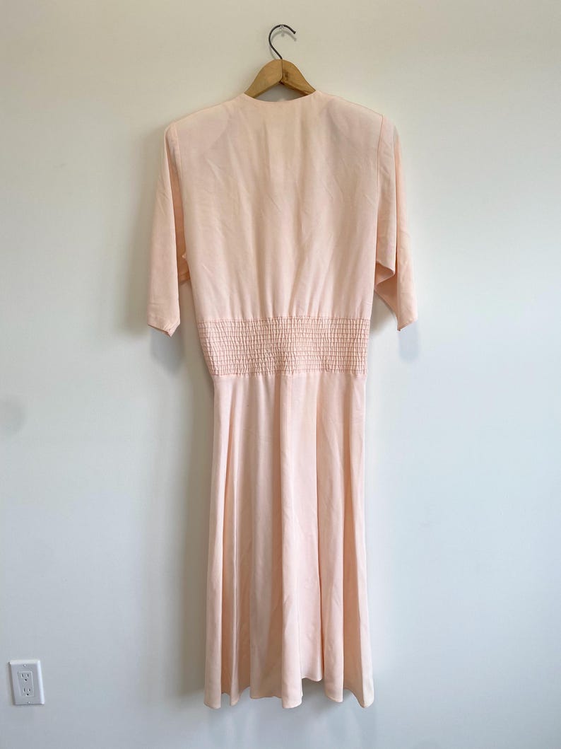 May include: A pale peach-colored midi dress featuring a V-neckline, elbow-length sleeves, and a wide smocked elasticized waist panel. The garment has a smooth, flowing skirt that falls below the knees and is displayed on a wooden hanger.