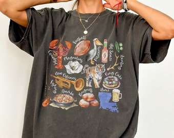 Puede incluir: Una camiseta gris oscuro con un estampado gráfico de varios iconos con temática de Luisiana, incluyendo un pelícano, un balón de fútbol, ​​un cangrejo de río, un tigre, un pastel de reyes, una trompeta y el estado de Luisiana. El texto de la camiseta incluye "Football", "Pelicans", "Tigers", "Hunting", "Hot Sauce", "King Cake", "New Orleans", "Canistys", "Magnolias", "Fishing", "Beignets", "Home", y "Jumbo".