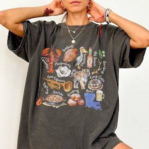 Puede incluir: Una camiseta gris oscuro con un estampado gráfico de varios iconos con temática de Luisiana, incluyendo un pelícano, un balón de fútbol, ​​un cangrejo de río, un tigre, un pastel de reyes, una trompeta y el estado de Luisiana. El texto de la camiseta incluye "Football", "Pelicans", "Tigers", "Hunting", "Hot Sauce", "King Cake", "New Orleans", "Canistys", "Magnolias", "Fishing", "Beignets", "Home", y "Jumbo".