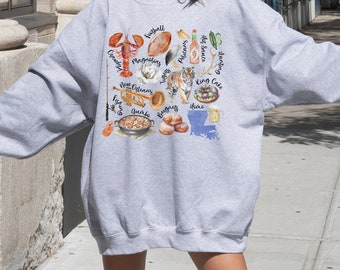 Puede incluir: Una sudadera gris con un estampado gráfico de varios artículos con temática de Luisiana, incluyendo un cangrejo de río, un balón de fútbol, un tigre, un pastel de rey y una cerveza. El texto "Sport Grey - 2XL" está impreso debajo de la imagen.