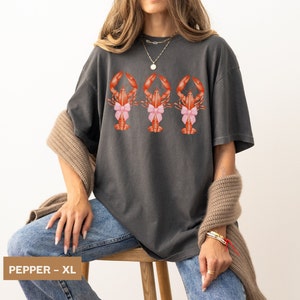 Pu&ograve; includere: Una maglietta oversize color antracite con una grafica di tre aragoste che indossano fiocchi rosa. Il testo "PEPPER - XL" &egrave; visibile nell'angolo in basso a sinistra dell'immagine.