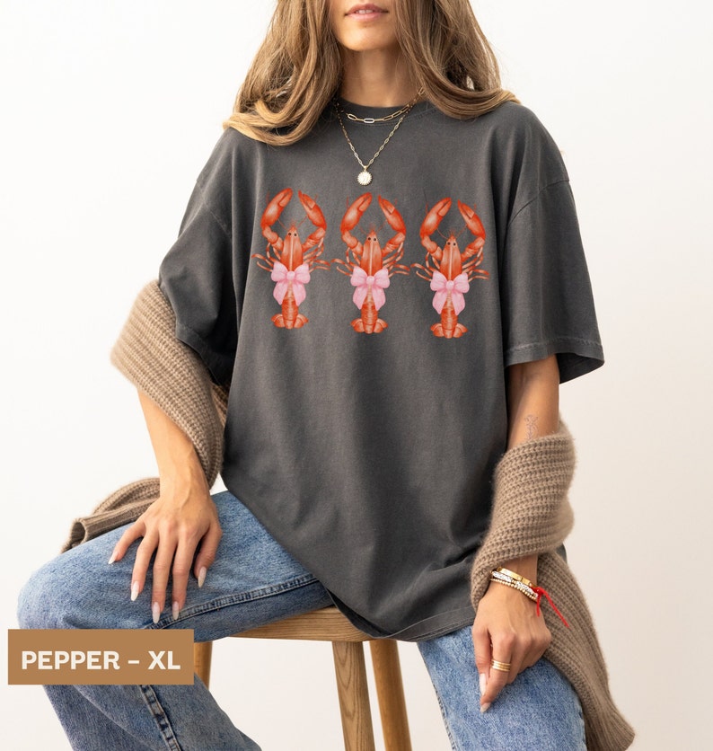Pu&ograve; includere: Una maglietta oversize color antracite con una grafica di tre aragoste che indossano fiocchi rosa. Il testo "PEPPER - XL" &egrave; visibile nell'angolo in basso a sinistra dell'immagine.