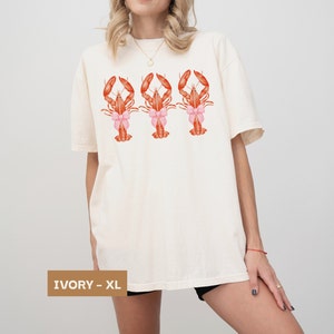 Pu&ograve; includere: Una maglietta bianca oversize con una grafica di tre aragoste rosse che indossano fiocchi rosa. La maglietta &egrave; disponibile in avorio e taglia XL.