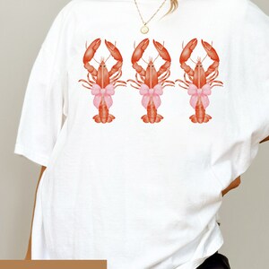 Pu&ograve; includere: Maglietta bianca oversize con una grafica di tre aragoste rosse che indossano fiocchi rosa. Il testo "WHITE - 2XL" &egrave; stampato nell'angolo in basso a sinistra della maglietta.