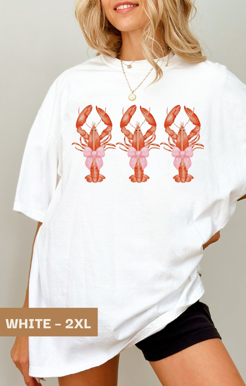 Pu&ograve; includere: Maglietta bianca oversize con una grafica di tre aragoste rosse che indossano fiocchi rosa. Il testo "WHITE - 2XL" &egrave; stampato nell'angolo in basso a sinistra della maglietta.