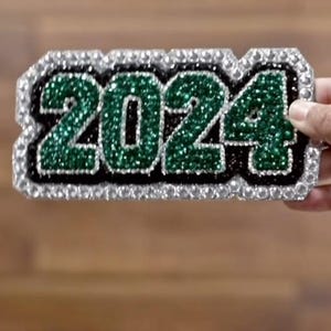 May include: A sparkly, rectangular patch with the year "2024" in green glitter. The numbers are outlined in black and surrounded by a border of silver rhinestones. The patch is held against a wooden background.
