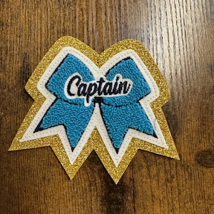May include: A blue and white bow with gold glitter trim and the word "Captain" in black lettering.