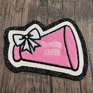 May include: A pink and white felt megaphone with a white bow and the text "Varsity Cheer" in white embroidery. The megaphone is surrounded by a black glitter border.