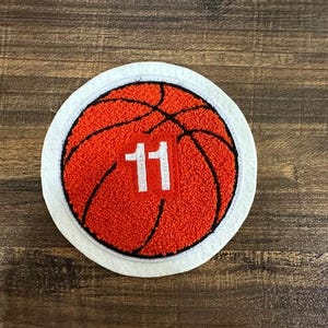 Puede incluir: Círculo de fieltro blanco con un balón de baloncesto bordado en rojo y negro con el número 11 en un cuadrado blanco.