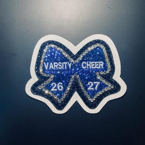 Puede incluir: Un parche azul y blanco con forma de lazo Varsity Cheer. El lazo está adornado con pedrería y presenta las palabras "VARSITY CHEER" y los números "26" y "27". El parche tiene un borde blanco sobre un fondo oscuro.