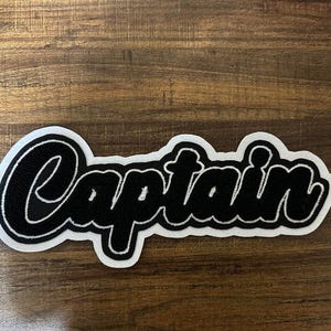 Puede incluir: Parche de fieltro blanco con letras bordadas en negro que dicen "Captain".