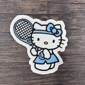 May include: White embroidered patch of Hello Kitty playing tennis. She is wearing a blue skirt and headband with a blue bow. She is holding a tennis racket and a tennis ball.