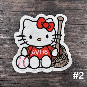 Könnte beinhalten: Weißer und roter gestickter Bügelaufkleber mit Hello Kitty, die ein rotes Shirt mit dem Text "AVHS" trägt und einen Baseballschläger und einen Baseball hält.