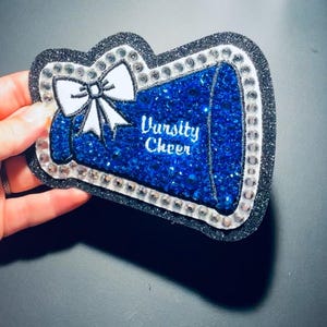 以下が含まれることがあります： 青と銀色のVarsity Cheerメガホン型の装飾品。メガホンは青いラインストーンで埋められ、銀色のラインストーンと黒いグリッターで縁取られています。上部には白いリボンがあり、「Varsity Cheer」の文字が白いスクリプトで書かれています。