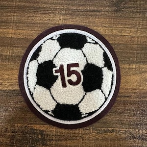 May include: A burgundy felt patch with a white stitched border, featuring a black and white soccer ball with the number 15 in maroon.