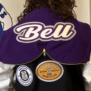Può includere: Giacca da wrestling viola e nera con una toppa "Bell" bianca e argentata sul retro. La giacca ha due toppe sul davanti, una dorata con "Masters Meet Southern Section 2024 Wrestling" e l'altra nera con "Wrestling Southern Section Championships 2024".