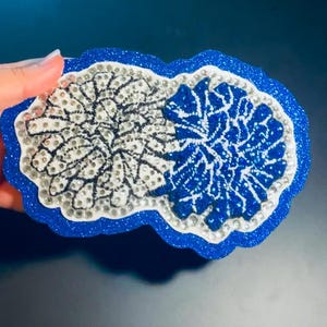 May include: A decorative coaster with a blue glitter border and a white and blue floral design. The coaster is embellished with rhinestones and has a unique, cloud-like shape. The design is split into two halves.