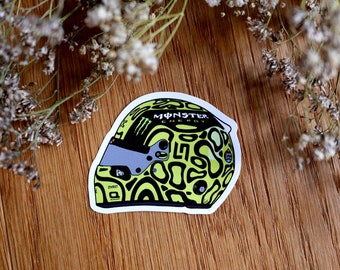 May include: A sticker of a racing helmet with a lime green and black pattern. The helmet has a grey visor and the Monster Energy logo. The sticker is on a wooden surface with dried flowers.