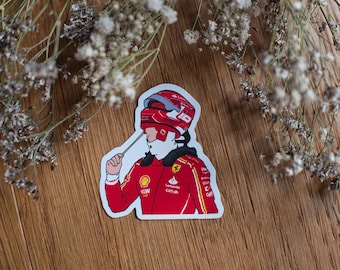 May include: A red and white racing driver sticker with a helmet and a red racing suit. The driver is wearing a white helmet with a red stripe and a red racing suit with a white sponsor logo. The sticker is on a wooden background.