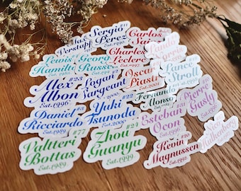 May include: A collection of colourful stickers featuring the names and numbers of Formula 1 drivers. Each sticker has a white background with a black outline and the driver's name and number in a stylised font. The stickers are arranged in a random pattern on a wooden surface.