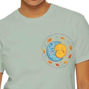 May include: A light green t-shirt featuring a circular graphic of a smiling sun and moon embracing. The design includes text that reads "MAY THE SUN BRING YOU NEW ENERGY BY DAY" and "MAY THE MOON SOFTLY RESTORE YOU BY NIGHT."