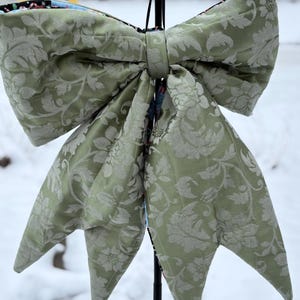 May include: A large, decorative bow in a soft green hue, featuring a detailed floral pattern. The bow is made of a textured fabric and has long, pointed tails. The bow is likely used for holiday or event decorations.
