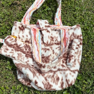 May include: A faux fur tote bag with a brown and white spotted pattern. The bag has a rectangular shape with a flap closure and a zippered pocket. The handles and trim feature a colorful, abstract design.