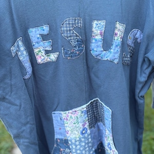 May include: A blue hoodie with the word "JESUS" appliqued across the front in patchwork fabric. The patchwork includes floral and plaid patterns in shades of blue. A matching patchwork pocket is sewn on the front.