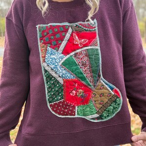 May include: A burgundy sweatshirt featuring a patchwork Christmas stocking design. The stocking is composed of various fabric patterns in red, green, blue, and brown, with a butterfly accent. The sweatshirt has a crew neck and long sleeves.