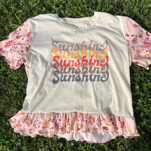 May include: Cream-colored t-shirt with the word "Sunshine" in a stacked, colorful design. The sleeves are a floral print, and the bottom has a ruffled floral trim. The shirt is laying on a bed of green grass.