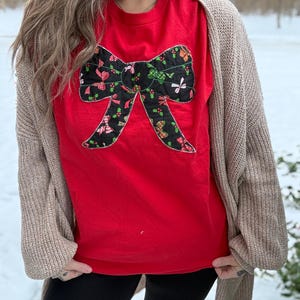 May include: A red t-shirt featuring a large, decorative bow design. The bow is black with a pattern of red, green, and white holiday-themed elements. A beige cardigan is worn over the t-shirt, and black leggings are visible.