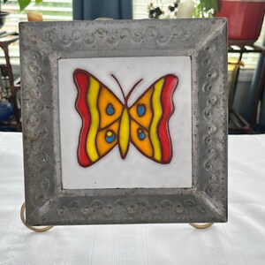 May include: A metal framed tile with a colorful butterfly design. The butterfly is yellow, red, and blue with black outlines. The frame is silver with a decorative border.