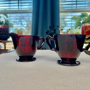 Könnte beinhalten: Ein Set aus zwei rubinroten Glaskrügen für Sahne und Zucker mit Griffen. Die Krüge haben ein klassisches Design und sind perfekt zum Servieren von Kaffee oder Tee.