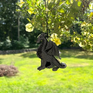 May include: A silver-colored dragon charm hanging from a black string. The dragon is in a sitting position with its wings spread out. The charm is hanging from a tree branch with yellow flowers.