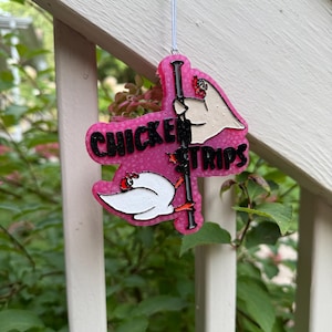 May include: A pink, textured, hanging ornament featuring two white chickens with red combs and orange feet. One chicken is positioned at the top and one at the bottom of a black vertical pole. The black text reads CHICKEN STRIPS across the center.