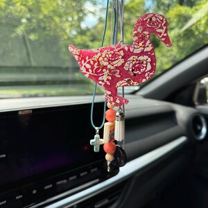 May include: A hanging car ornament shaped like a duck, featuring a bright pink base with a white floral pattern and gold glitter accents. It is suspended by cords adorned with a turquoise cross charm, round peach beads, a white tassel, and a dark stone.