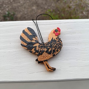 May include: A decorative chicken ornament with a black string for hanging. The chicken has a tan body with black markings, a red comb, and gold legs. The ornament is made of a textured material.