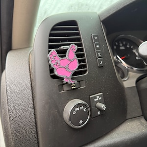 May include: A pink chicken-shaped car air freshener hanging from the air vent of a car's dashboard.