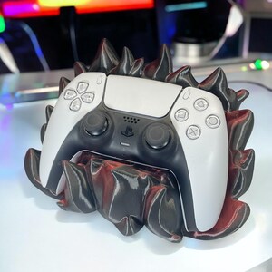 Könnte beinhalten: Ein weiß-schwarzer PlayStation 5-Controller liegt in einem speziell angefertigten Halter. Der Halter ist schwarz mit roten Akzenten und einem stacheligen, strukturierten Design. Der Controller zeigt das PlayStation-Logo und die Tasten.