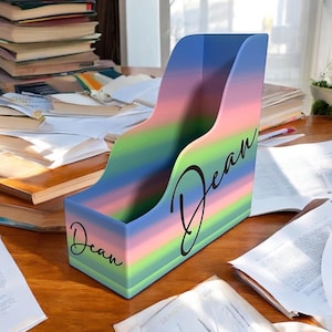 Könnte beinhalten: Ein vertikaler Zeitschriftenhalter mit einem horizontalen Farbverlauf aus blauen, rosa und grünen Streifen. Der Name Dean ist in schwarzer Schreibschrift sowohl auf der Vorderseite als auch auf der Seitenwand des aufrechten Behälters gedruckt.
